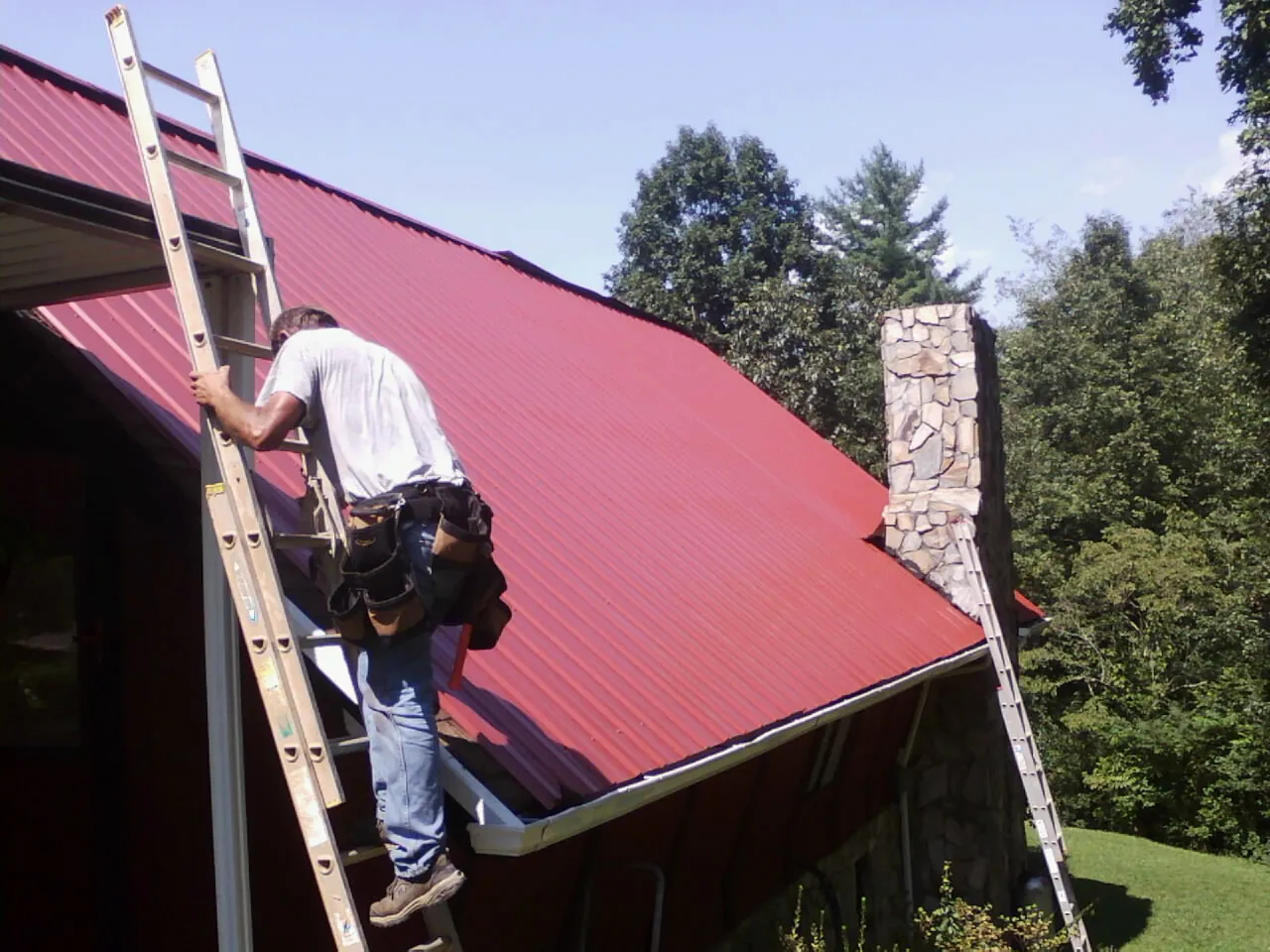 Roofer on a ladder beside a red metal roof and stone chimney — Soffit & Fascia Repair by Roofing Brothers in Shorewood